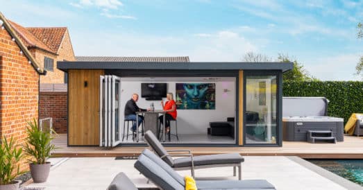 Front view of garden bar with slidefold doors opena nd customers enjoying a drink on inside table. Grey rattan sun loungers in the foreground. contemporary garden room by a pool